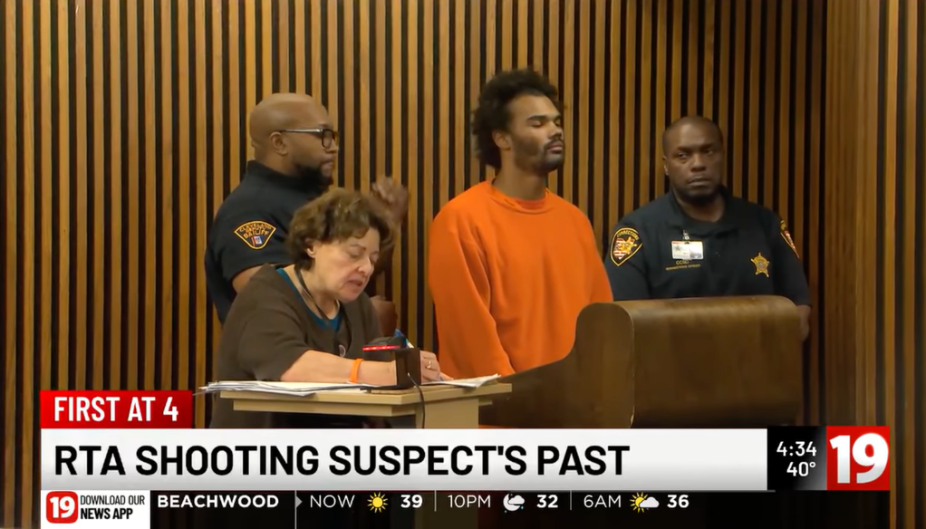 A man in an orange jumpsuit stands in court with two bailiffs behind him and a woman taking notes at a table in front of him, with news graphics overlaid.